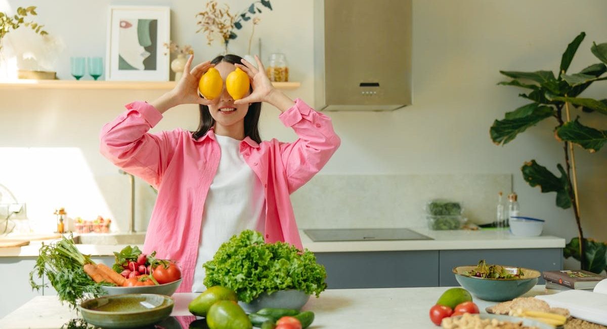A woman playfully holding lemons over her eyes in a bright, lively kitchen setting filled with fresh vegetables.