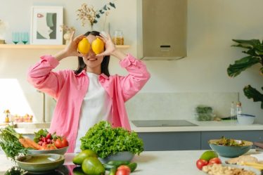 A woman playfully holding lemons over her eyes in a bright, lively kitchen setting filled with fresh vegetables.