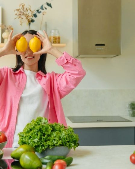 A woman playfully holding lemons over her eyes in a bright, lively kitchen setting filled with fresh vegetables.